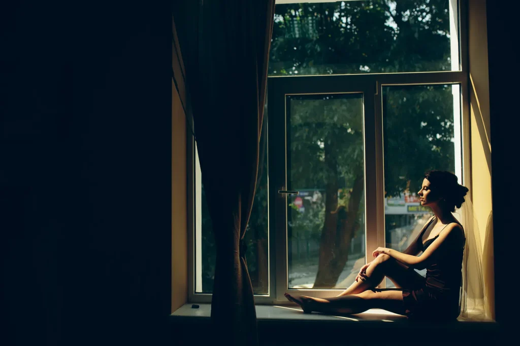 A woman sitting alone on a windowsill in a dim room, gazing out of the window