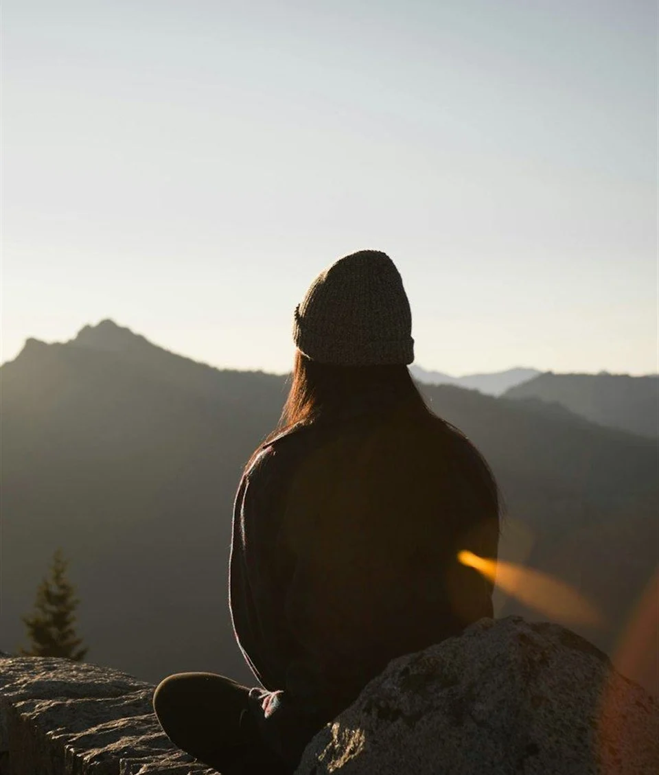 A person sitting on a rock at the edge of a ridge, looking out over distant mountains