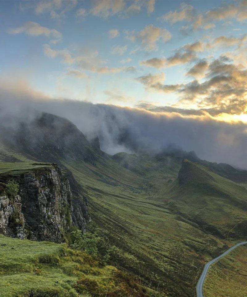 A winding road leading through green hills towards a bright, cloud-lit horizon