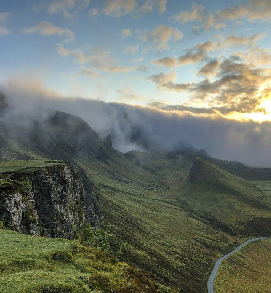 Green hills and a winding road beneath soft clouds and evening light