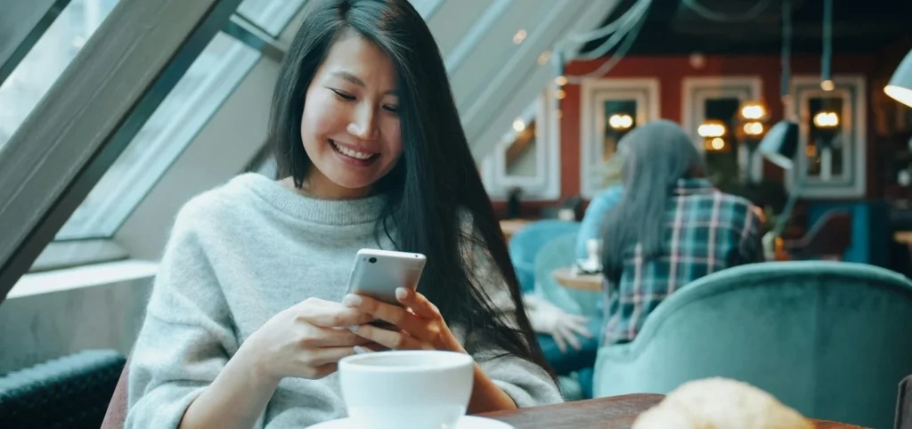 A smiling woman looking at her phone over a cup of coffee in a warm cafe, getting in touch