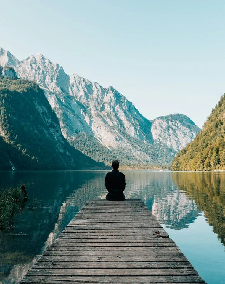 A person sitting calmly at the end of a wooden jetty, looking across a still mountain lake