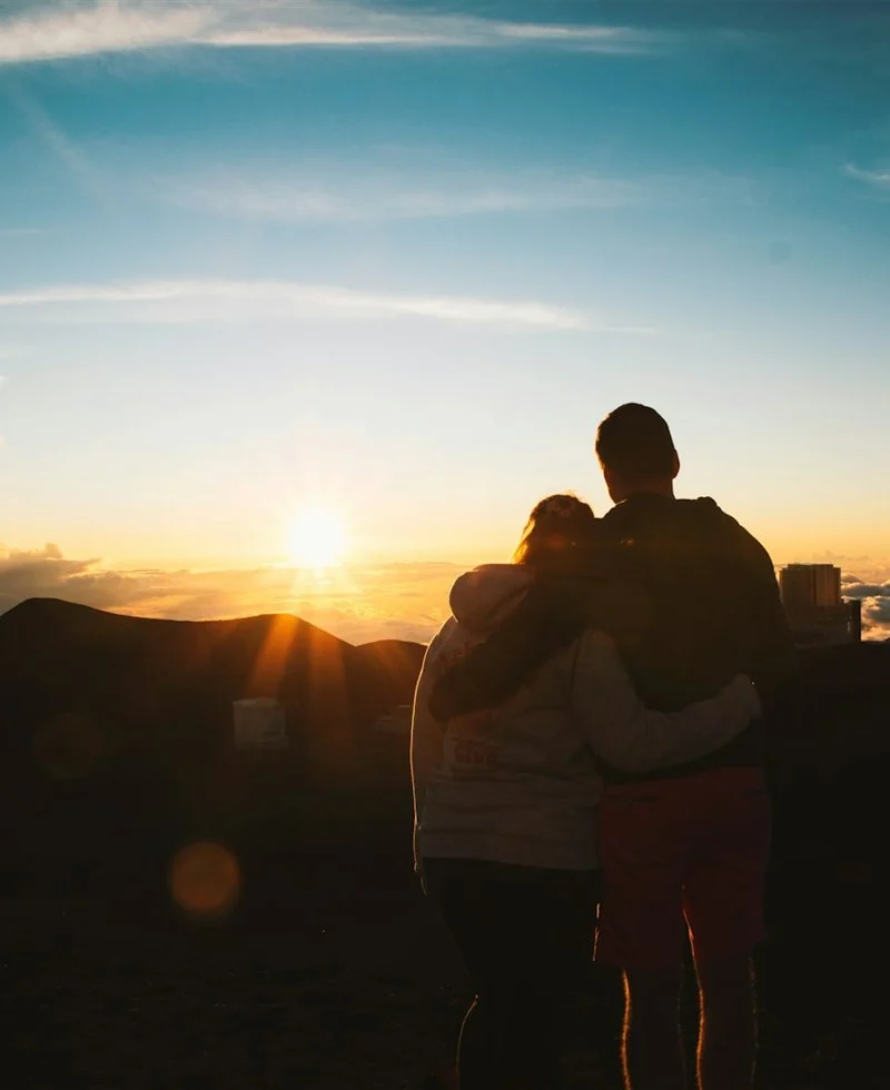 Two people standing close together in silhouette as the sun sets above the clouds