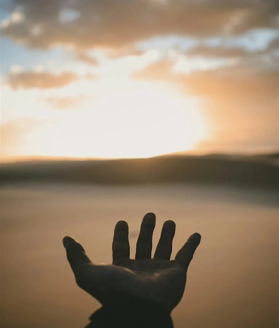 An open hand reaching up towards a soft, warm sunrise over still water